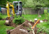Grävmaskin i trädgård gräver en rörgrav i gräsmattan.