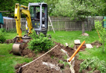 Grävmaskin i trädgård gräver en rörgrav i gräsmattan.