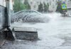 Regn rinner från ett stuprör under ett kraftigt regn. Bil kör genom en vattensamling i bakgrunden.