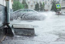 Regn rinner från ett stuprör under ett kraftigt regn. Bil kör genom en vattensamling i bakgrunden.