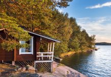 Enkel brun trästuga med vita fönsterfoder, på en klippa vid vatten. Skog runt om.