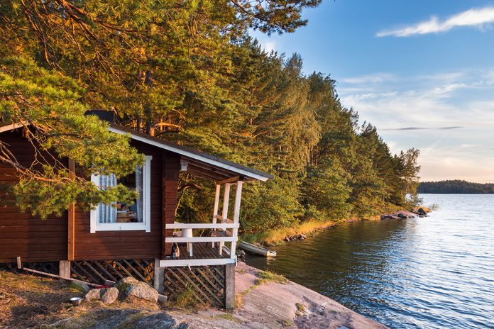 Enkel brun trästuga med vita fönsterfoder, på en klippa vid vatten. Skog runt om.