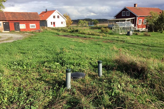 Två plaströr sticker upp ur marken, bredvid ett svart plastlock. Grönt gräs och ett vitt och ett rött hus i bakgrunden.