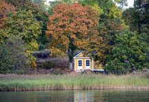 gult hus med vita knutar, omgivet av lövträd som börjar skifta i höstfärger. Längst fram mörkt vatten i en sjö och vass mellan sjön och huset.