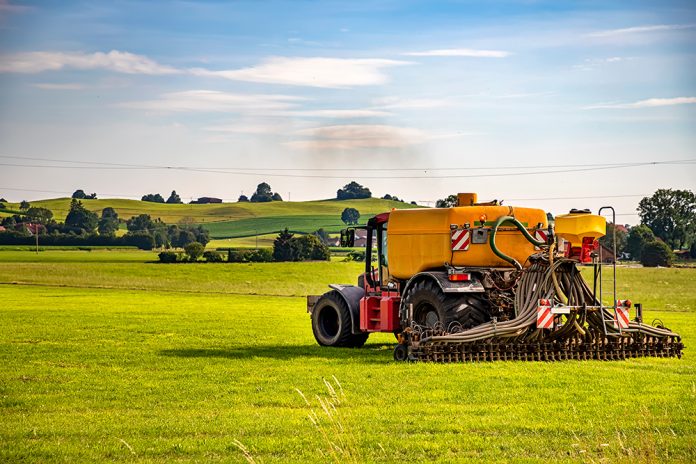 Traktor sprider flytgödsel på gräs. Gröna kullar i bakgrunden.