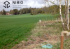 Grönt fält med en avloppsanläggning i åkerkanten, i förgrunden. NIBIO:s logotyp i övre vänstra hörnet.