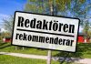 Skylt med texten redaktören rekommenderar. Bakom finns grönt gräs, en björk och röda stugor.